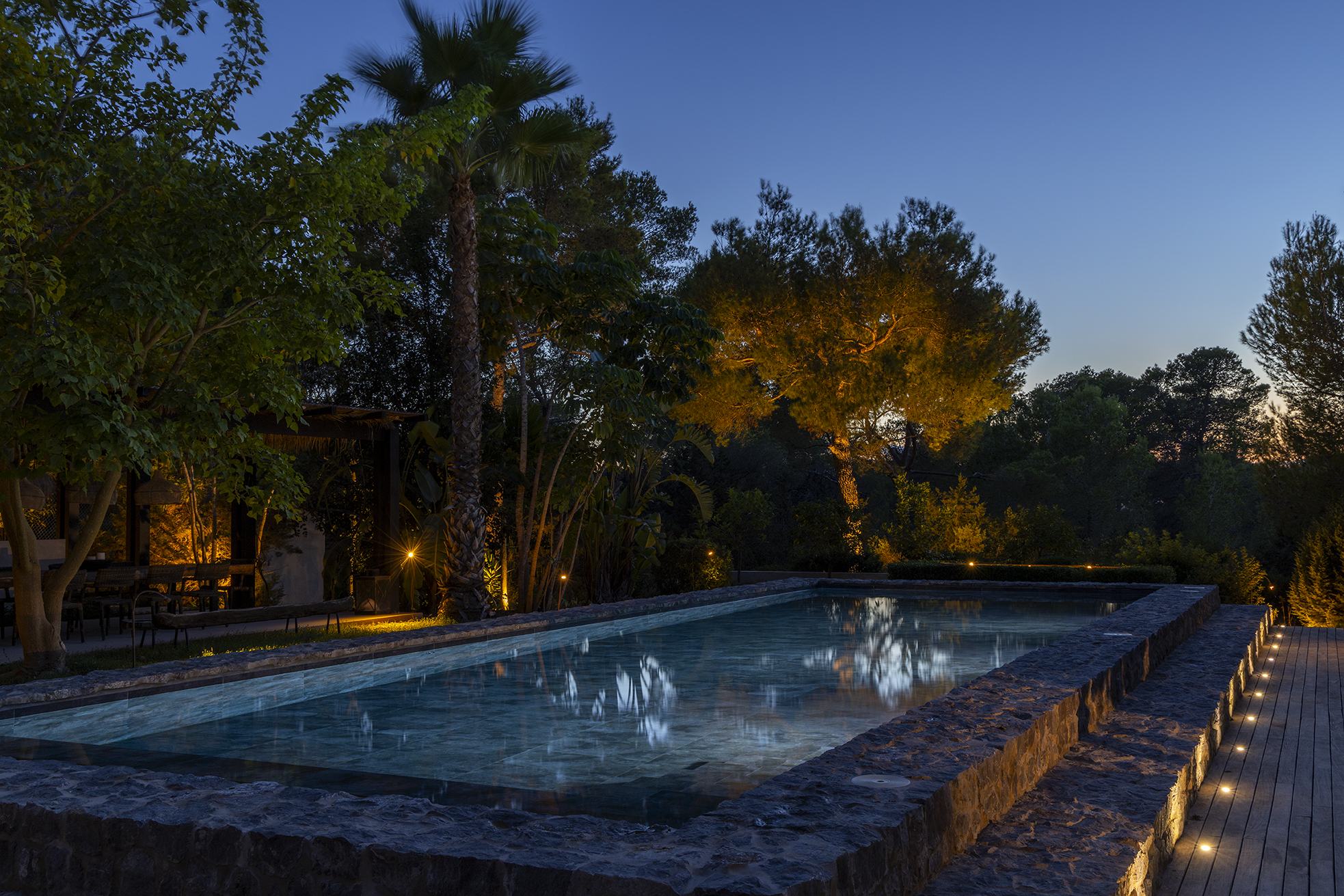 Can Serenico pool at dusk — stone-edged pool, palm and pines lit warm against a deep blue sky